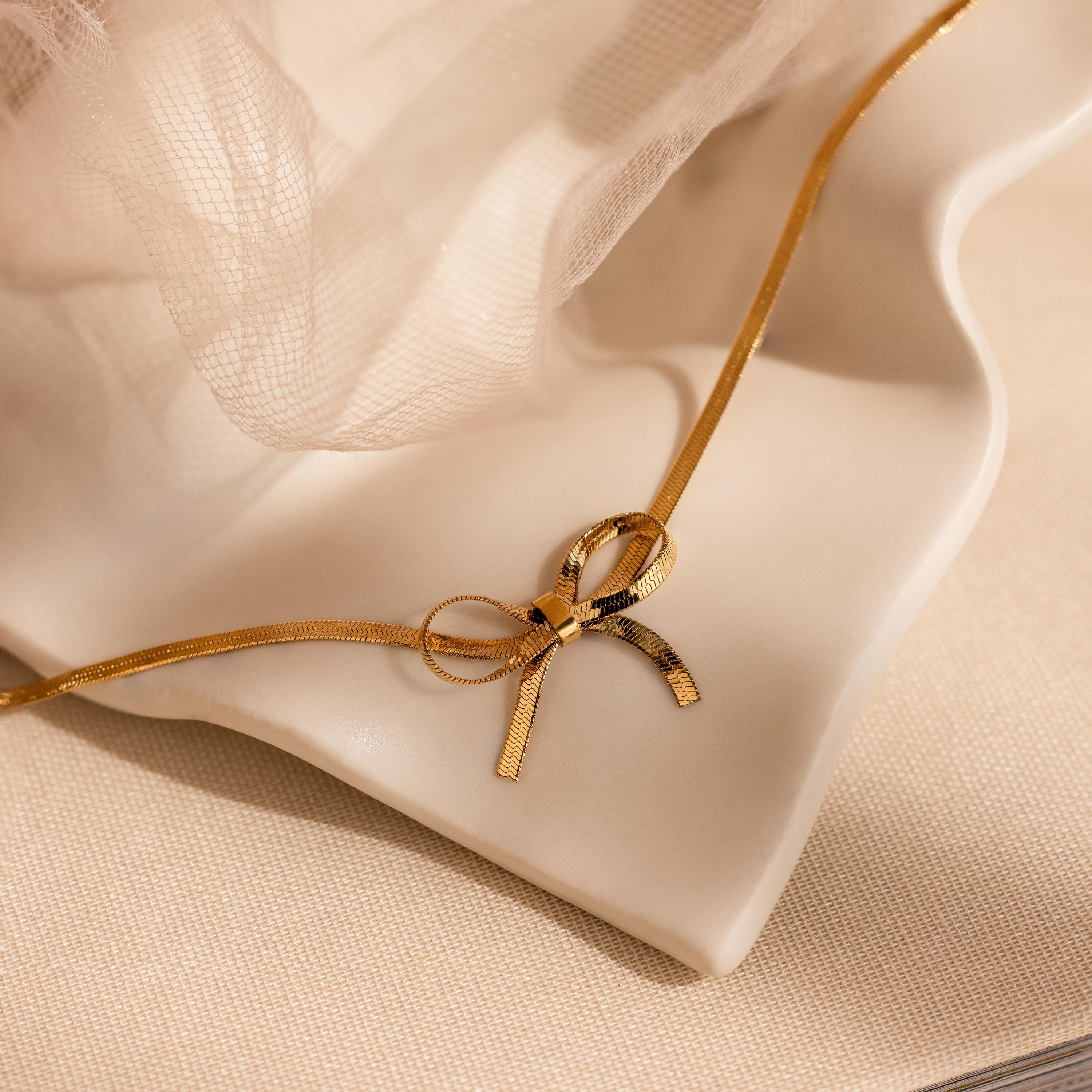 The Herringbone Bow Necklace in 18K Gold, styled in a coquette fashion, is showcased on a cream wavy ceramic dish with soft fabric in the background.