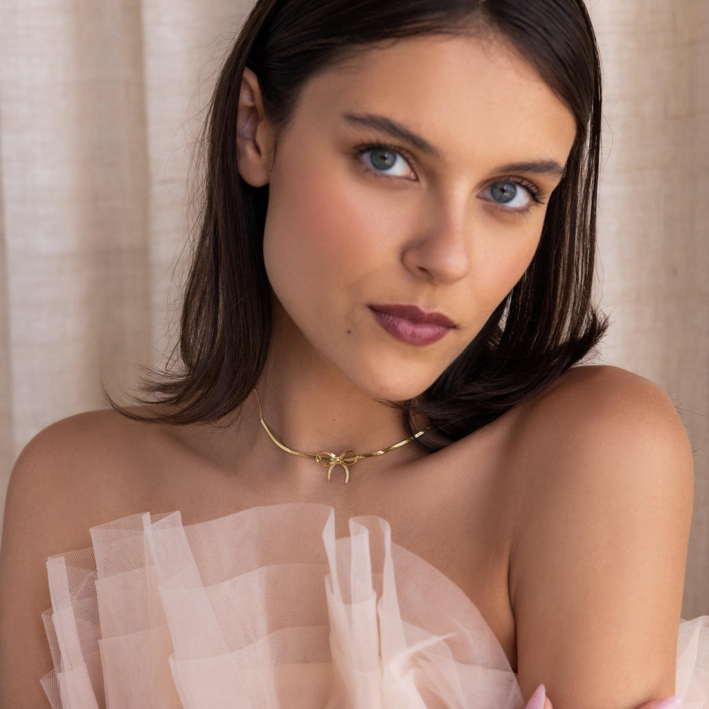 A woman with blue eyes and brown hair wears a sheer, ruffled top and the Herringbone Bow Necklace in 18K Gold, gazing at the camera in a coquette style.