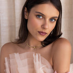 A woman with blue eyes and brown hair wears a sheer, ruffled top and the Herringbone Bow Necklace in 18K Gold, gazing at the camera in a coquette style.