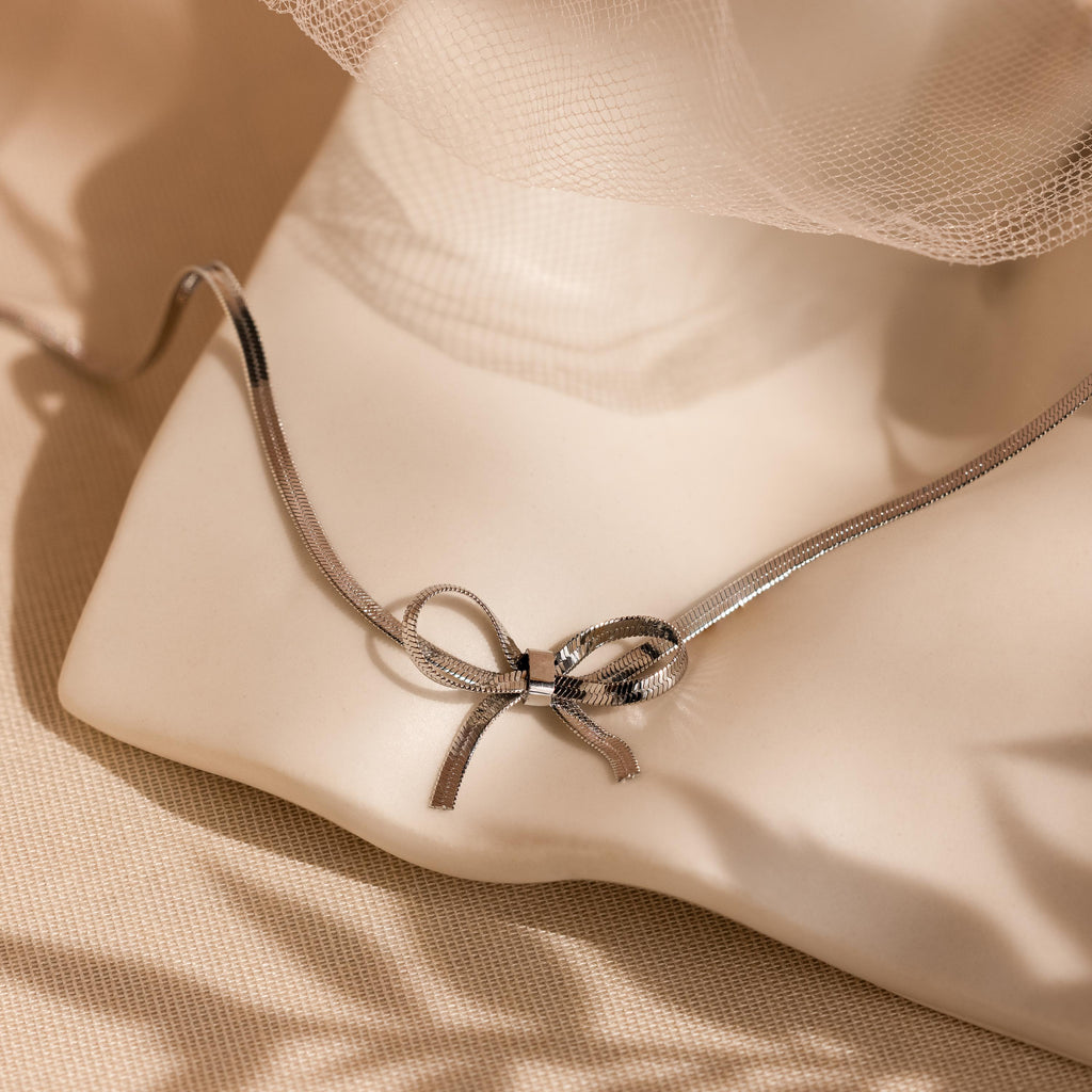 The Herringbone Bow Necklace in Sterling Silver rests on a white sculpture with soft beige fabric behind it—a charming accent for coquette style.
