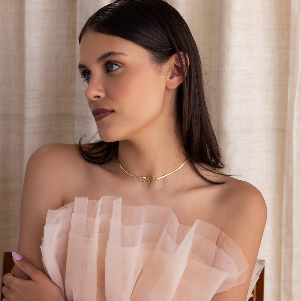 A woman with dark hair wears an off-shoulder, ruffled pale dress and the Herringbone Bow Necklace, capturing a soft coquette look as she gazes to the side.