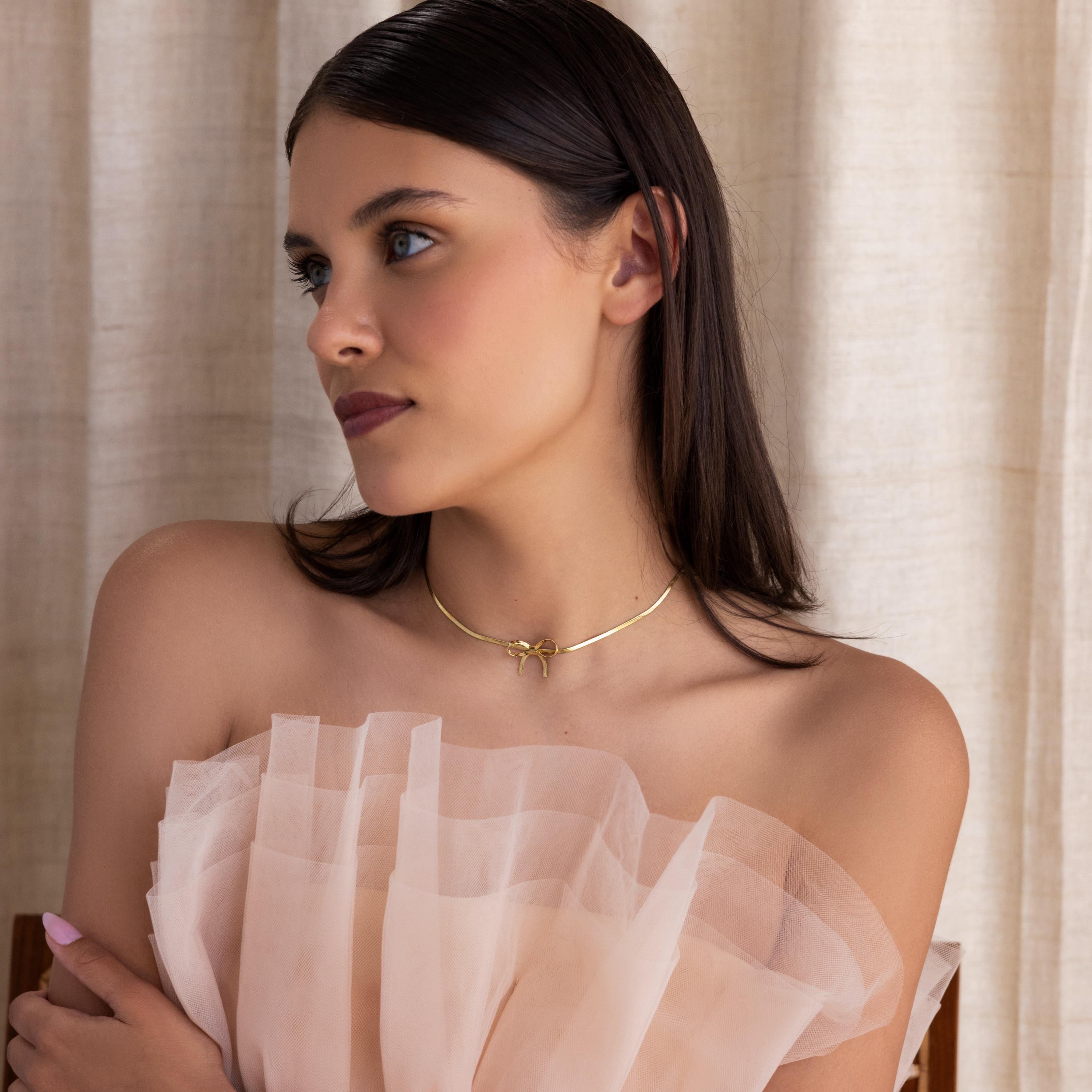 A woman with dark hair wears an off-shoulder, ruffled pale dress and the Herringbone Bow Necklace, capturing a soft coquette look as she gazes to the side.