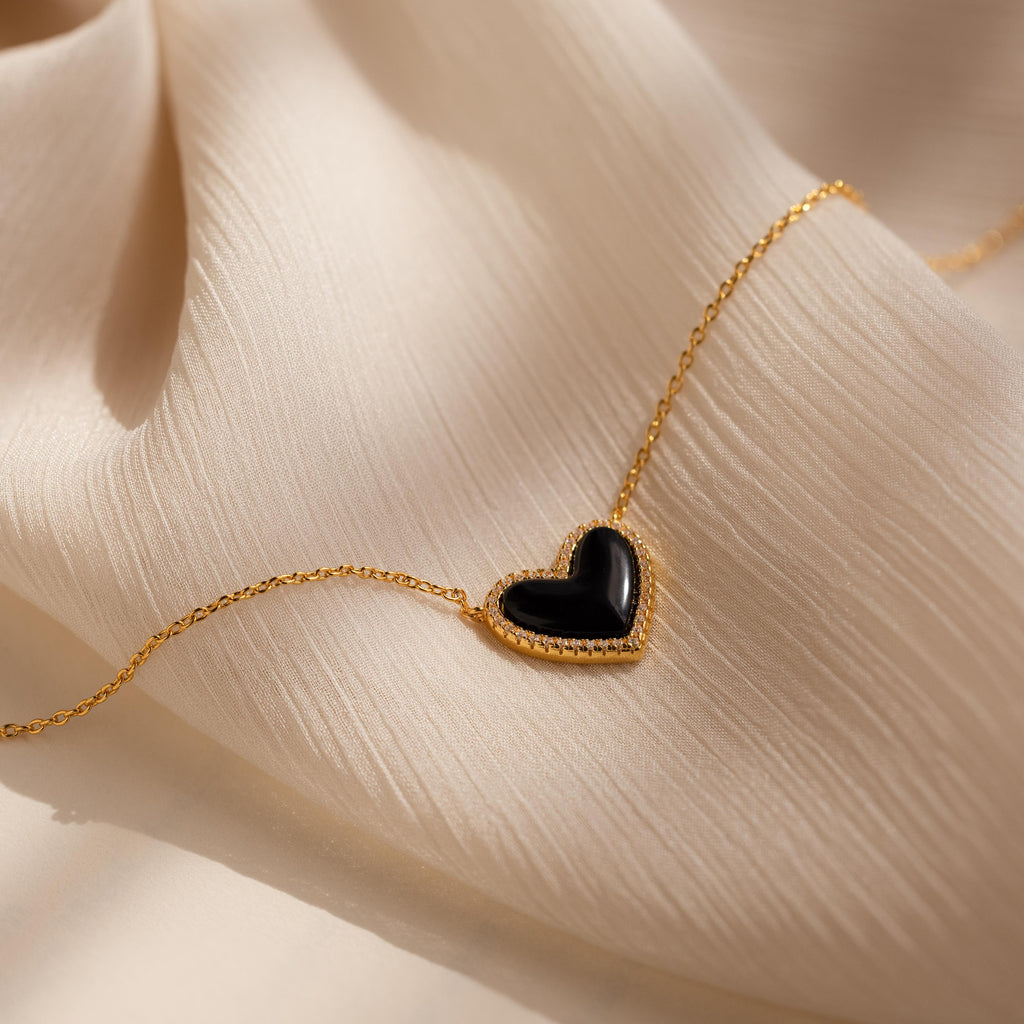 The Pave Onyx Puff Heart Necklace in 18K Gold, a coquette-inspired piece with a black onyx heart pendant, is shown resting on a soft beige fabric background.