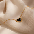 The Pave Onyx Puff Heart Necklace in 18K Gold, a coquette-inspired piece with a black onyx heart pendant, is shown resting on a soft beige fabric background.