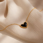 The Pave Onyx Puff Heart Necklace in 18K Gold, a coquette-inspired piece with a black onyx heart pendant, is shown resting on a soft beige fabric background.