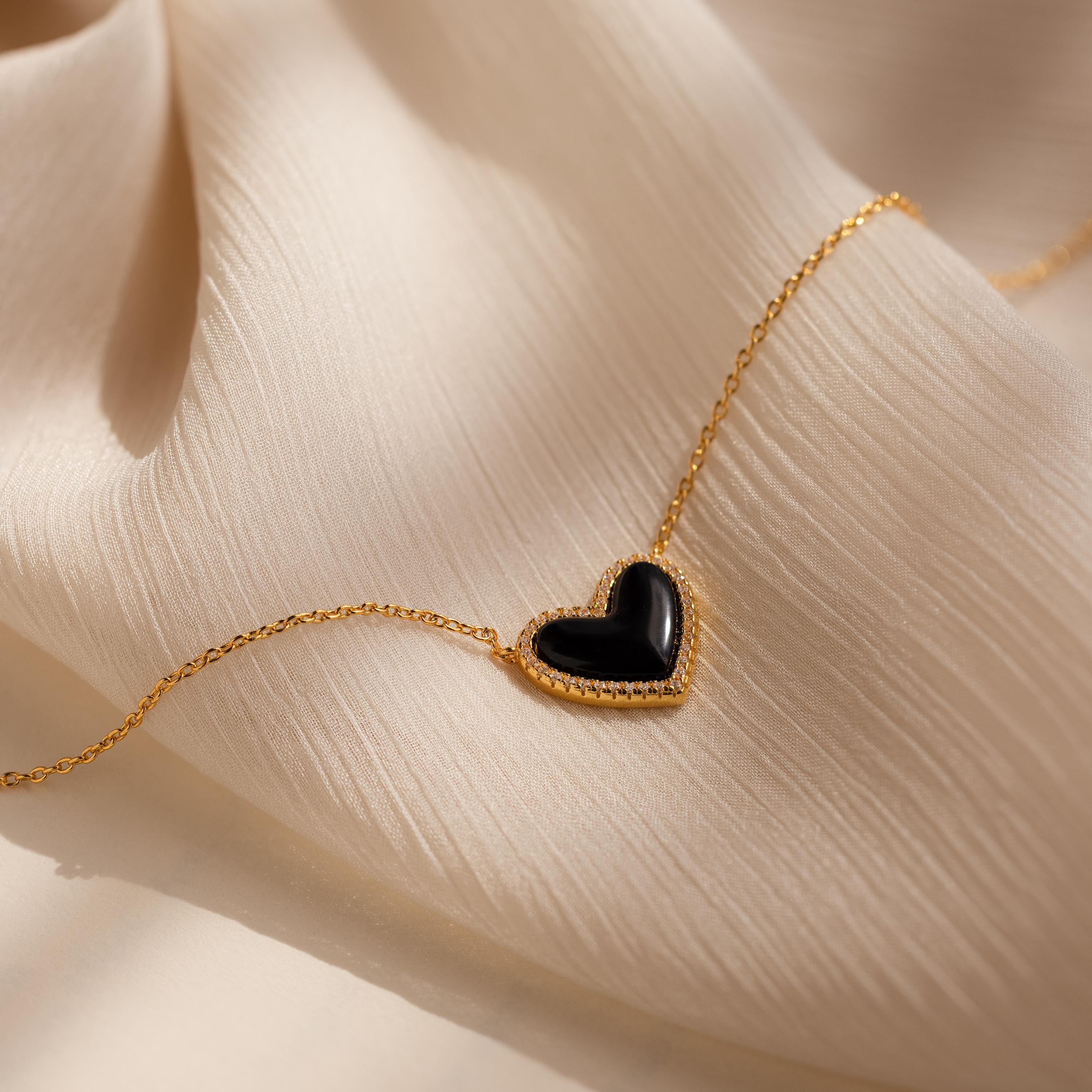 The Pave Onyx Puff Heart Necklace in 18K Gold, a coquette-inspired piece with a black onyx heart pendant, is shown resting on a soft beige fabric background.