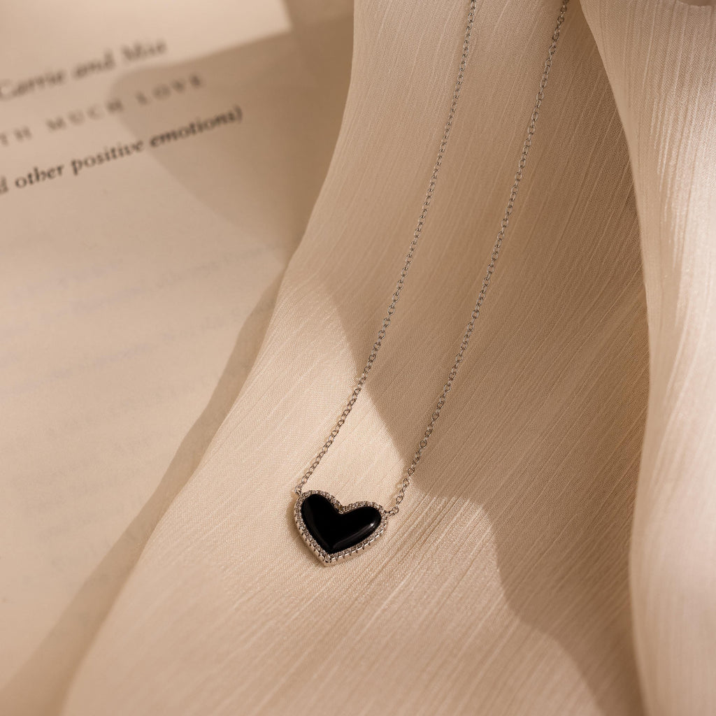 The Pave Onyx Puff Heart Necklace in Sterling Silver, featuring a black heart pendant, rests on cream-colored textured fabric and adds a touch of elegance to the scene.