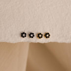 The Onyx Flower Studs set includes four small, flower-shaped flatback earrings—two silver and two gold—displayed on a textured beige surface. These elegant studs add subtle charm to any look.