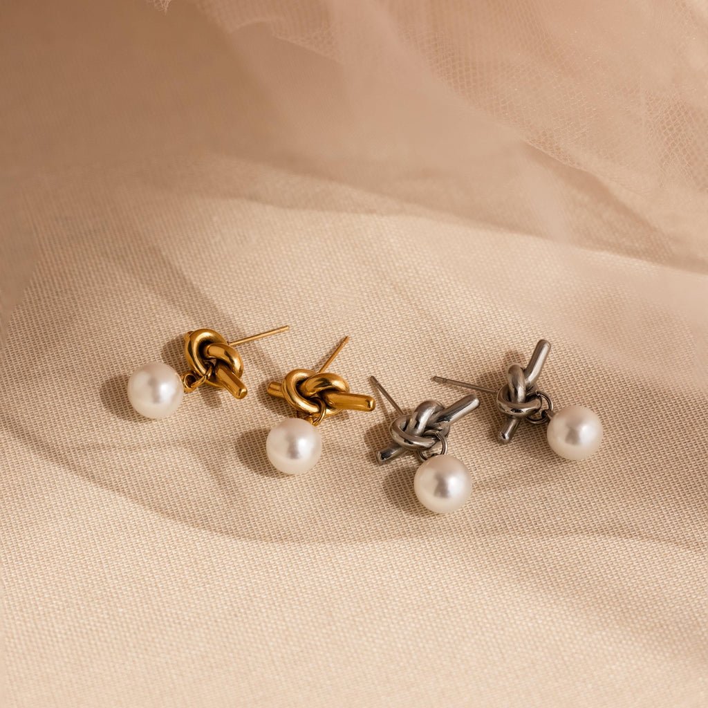 Two pairs of Love Knot Pearl Earrings, featuring pearl drops in gold and silver tones, are displayed on beige fabric—a timeless wedding accessory ideal for vintage-inspired jewelry lovers.