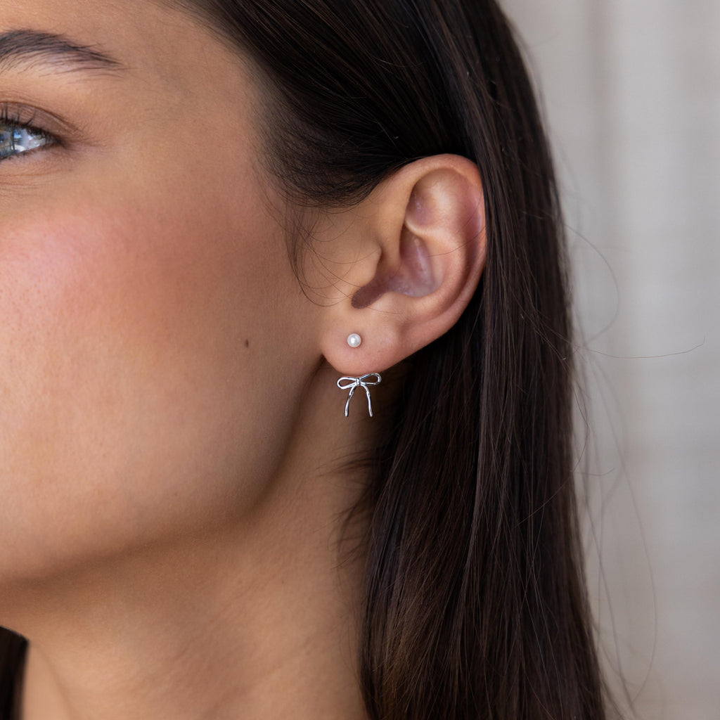 Close-up of a woman’s ear wearing the Pearl & Bow Ear Jackets in Sterling Silver, showcasing the elegant combination of a small pearl stud with a delicate silver bow accent.