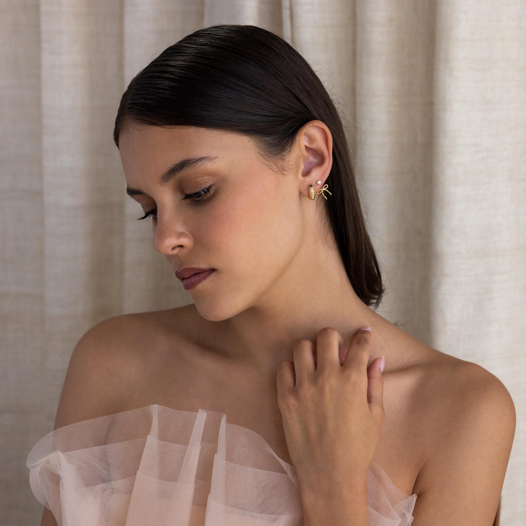 A woman with dark hair in an off-shoulder sheer dress looks down, softly touching her collarbone as her Pearl & Bow Ear Jackets elegantly catch the light.