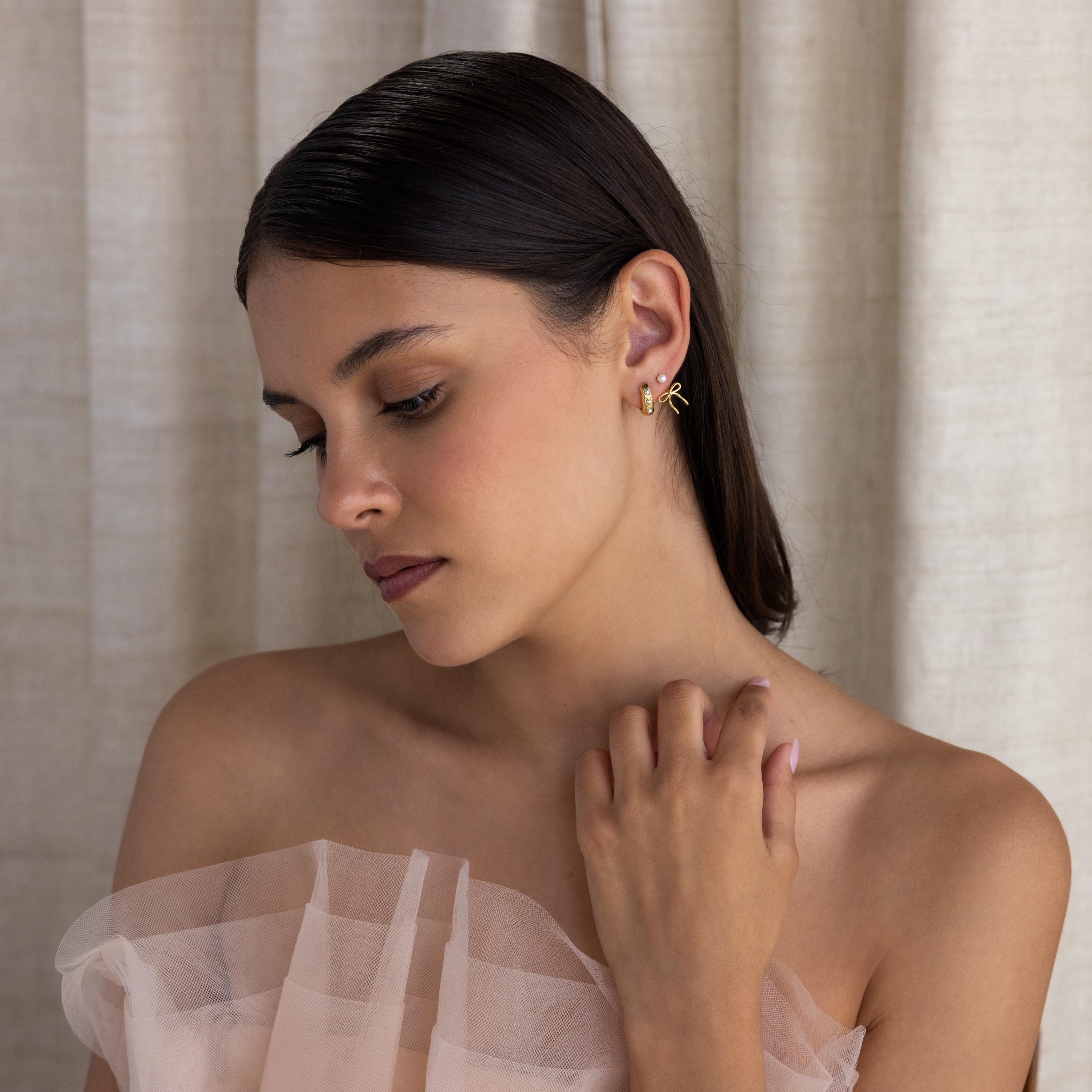 A woman with dark hair in an off-shoulder sheer dress looks down, softly touching her collarbone as her Pearl & Bow Ear Jackets elegantly catch the light.