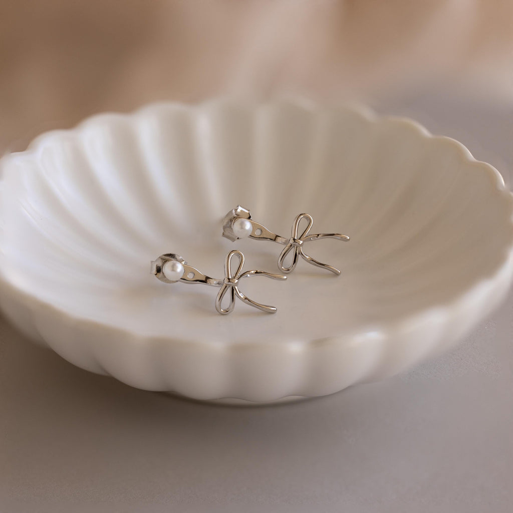 The Pearl & Bow Ear Jackets feature delicate bows and small pearls, elegantly displayed on a scalloped white ceramic dish.