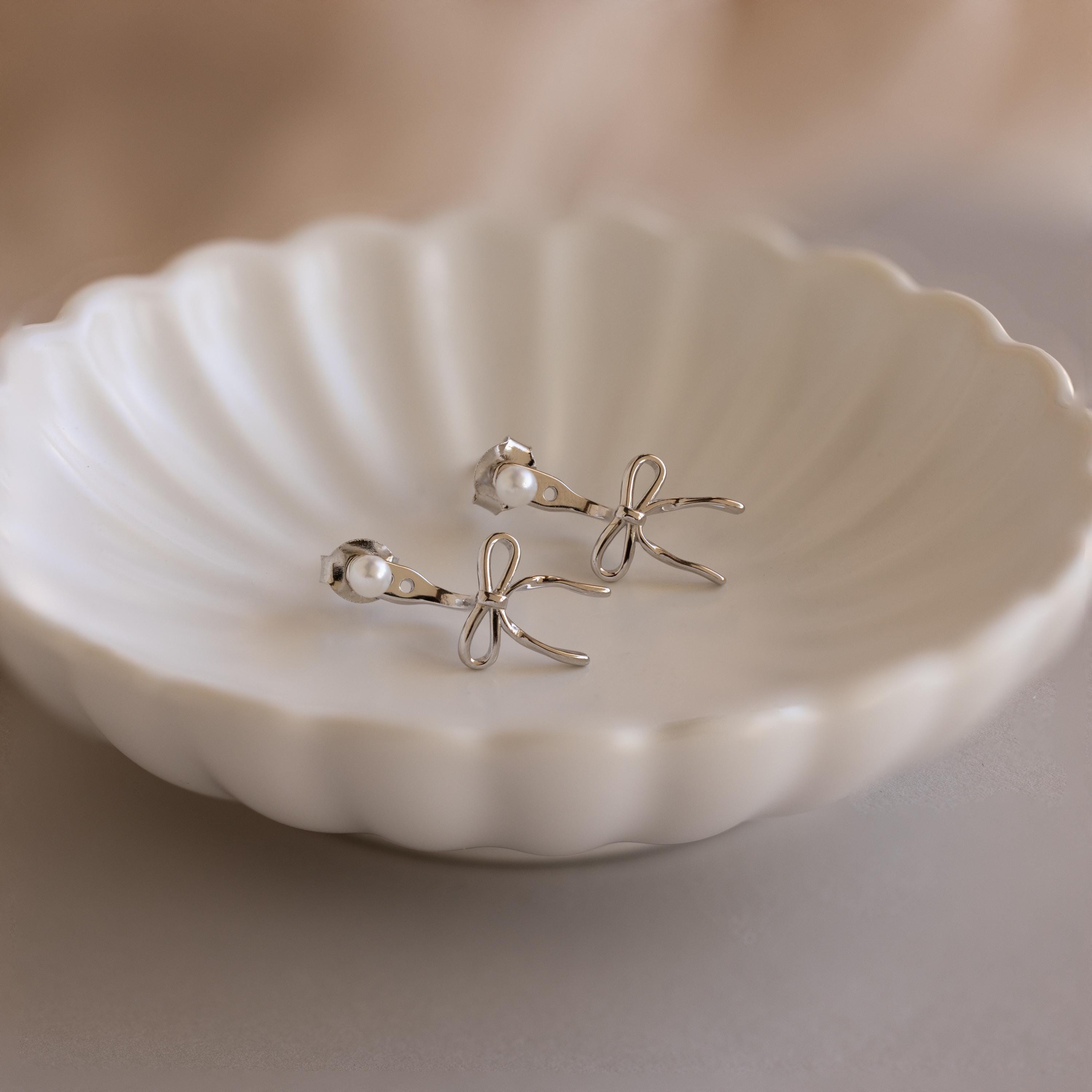 The Pearl & Bow Ear Jackets feature delicate bows and small pearls, elegantly displayed on a scalloped white ceramic dish.