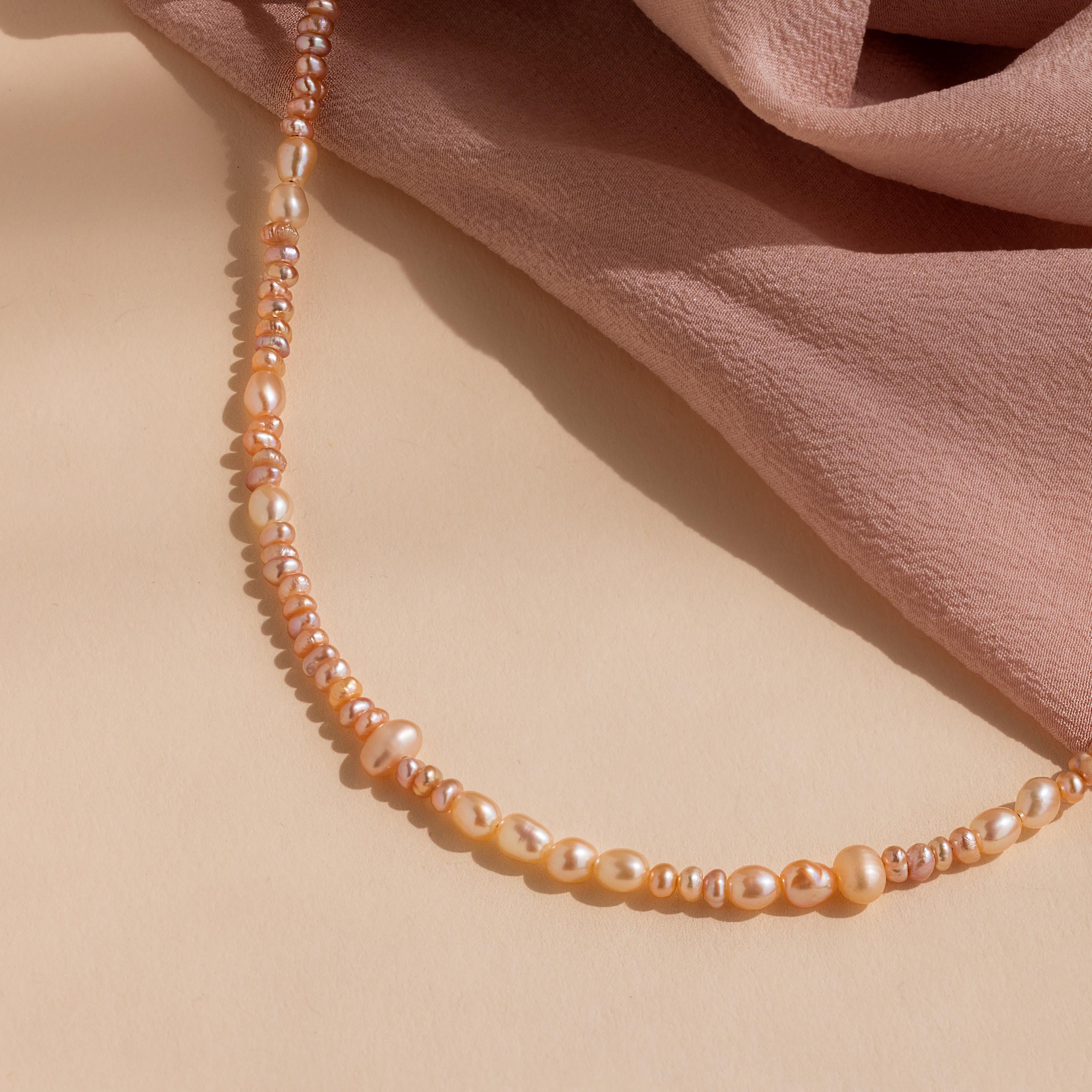 The Blush Pearl Beaded Necklace, showcasing a string of pink freshwater pearls, is displayed on a beige surface next to blush pink fabric.