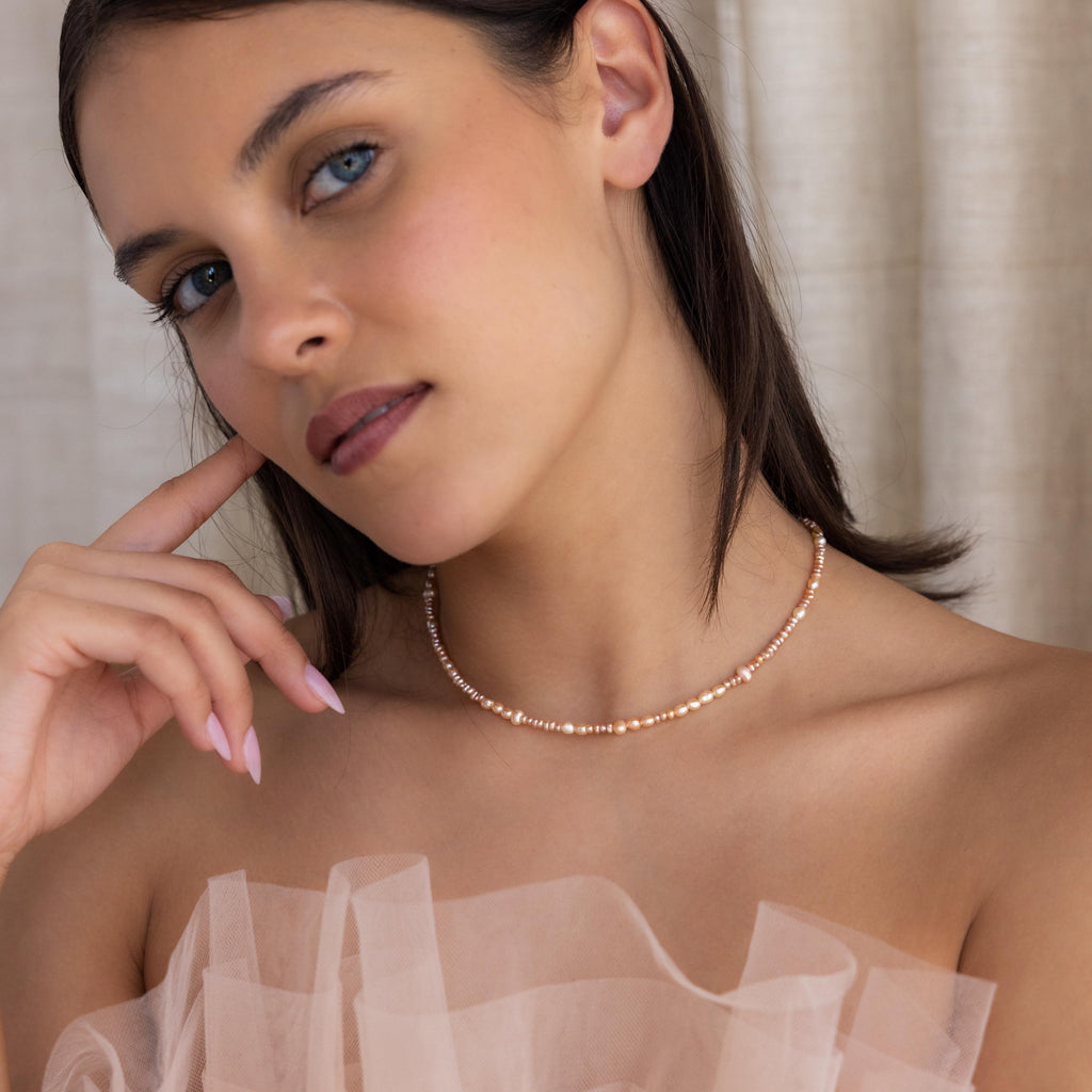 Blue-eyed woman wearing the Blush Pearl Beaded Necklace and a sheer, nude-colored top, posing with her hand near her face.