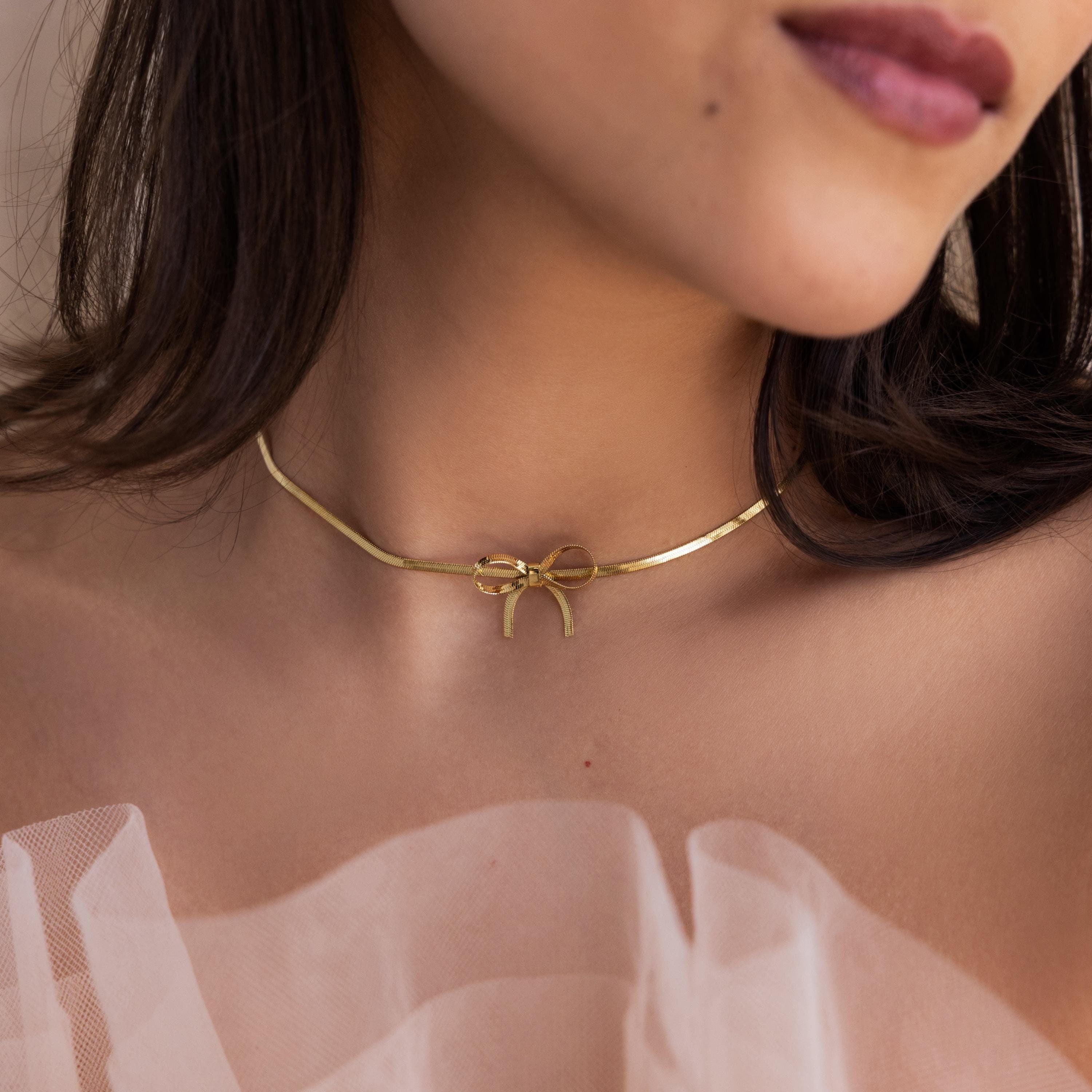 Wearing the Herringbone Bow Necklace layered with a gold choker and sheer light pink fabric, she achieves a dreamy coquette style.