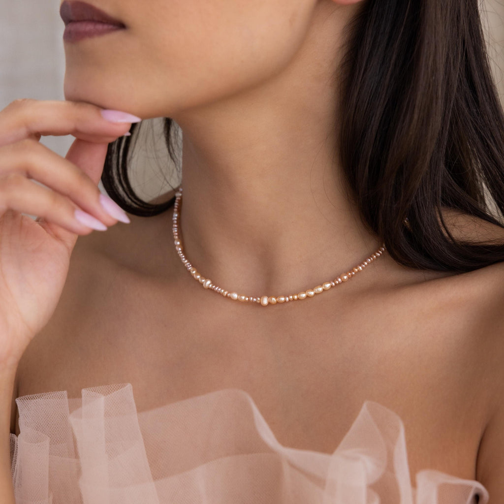 A woman thoughtfully touches her face while wearing a sheer nude top and the Blush Pearl Beaded Necklace.
