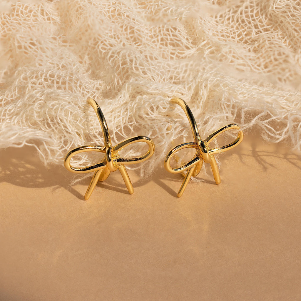 The Classic Ribbon Bow Huggies, gold bow-shaped earrings, are displayed on textured beige fabric—ideal for adding a feminine touch to any outfit.