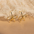 The Classic Ribbon Bow Huggies, gold bow-shaped earrings, are displayed on textured beige fabric—ideal for adding a feminine touch to any outfit.