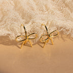 The Classic Ribbon Bow Huggies, gold bow-shaped earrings, are displayed on textured beige fabric—ideal for adding a feminine touch to any outfit.