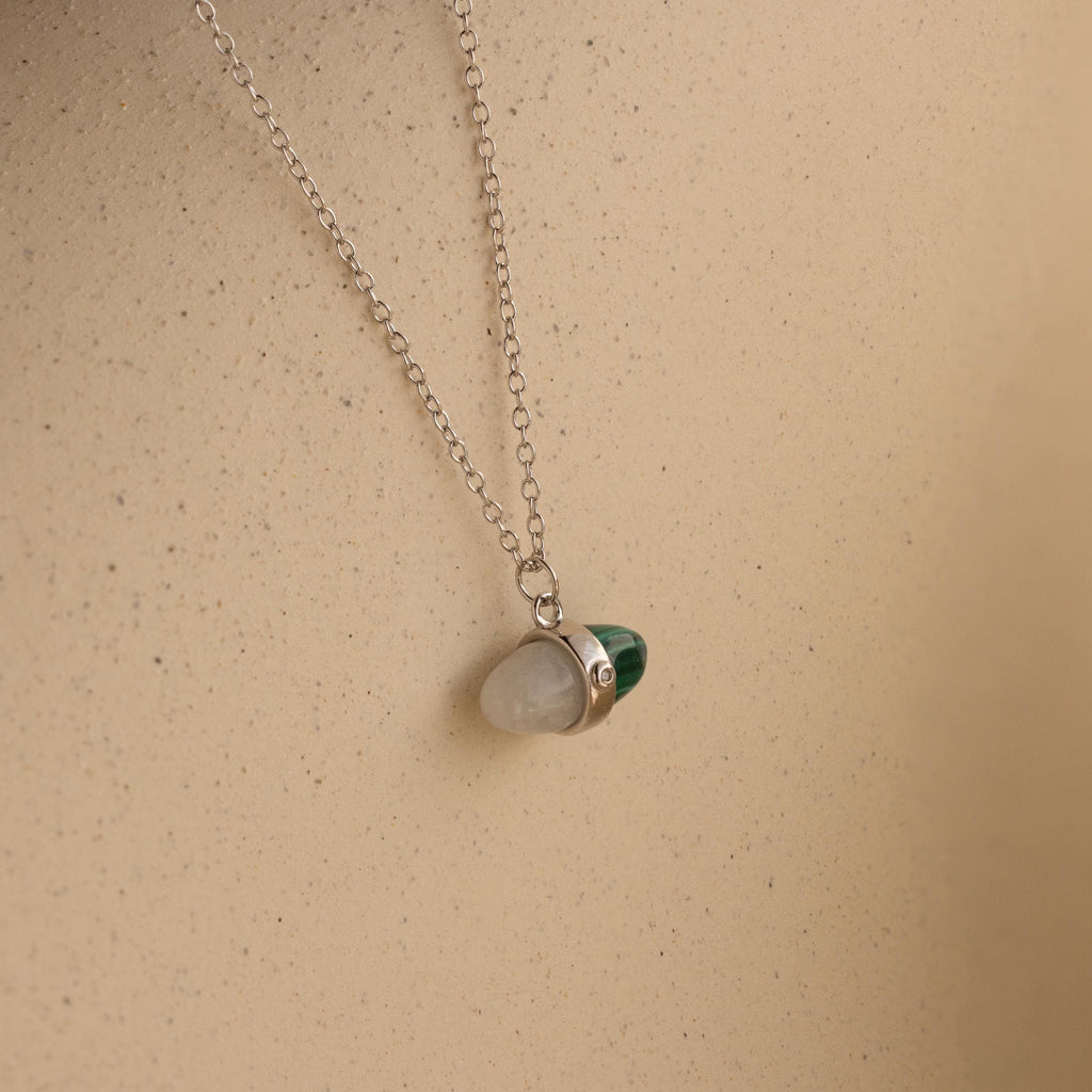 The Malachite & Moonstone Necklace features a silver chain with moonstone and malachite gemstone pendants, displayed on a beige background—ideal for those looking for healing gemstone jewelry.