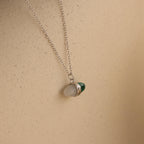 The Malachite & Moonstone Necklace features a silver chain with moonstone and malachite gemstone pendants, displayed on a beige background—ideal for those looking for healing gemstone jewelry.