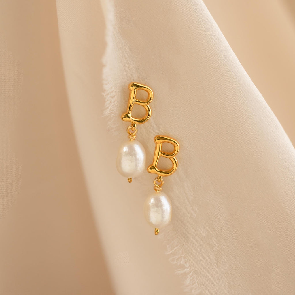 The Initial Pearl Drop Studs feature gold studs with letter B charms and dangling white pearls, displayed on a soft beige fabric background.