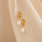 The Initial Pearl Drop Studs feature gold studs with letter B charms and dangling white pearls, displayed on a soft beige fabric background.