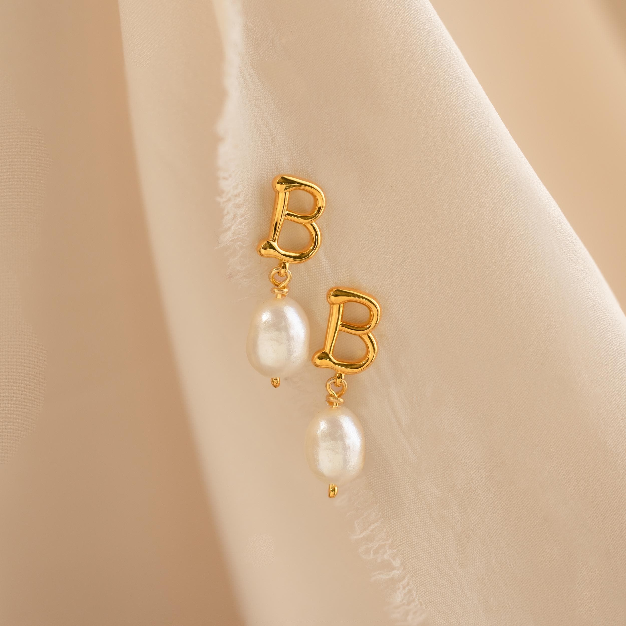 The Initial Pearl Drop Studs feature gold studs with letter B charms and dangling white pearls, displayed on a soft beige fabric background.