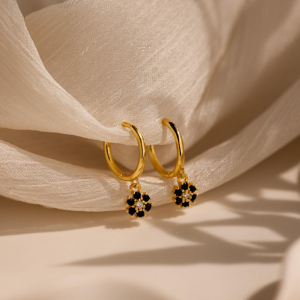 Onyx Flower Huggies: gold hoop earrings with small black and white floral charms, displayed on soft, sheer white fabric.