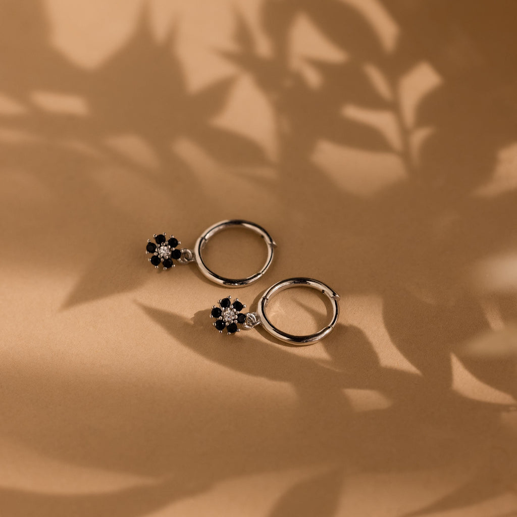 Two Onyx Flower Huggies, featuring silver hoops with black onyx flower charms, rest on a brown surface with leafy shadows, highlighting their elegant floral design.