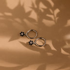 Two Onyx Flower Huggies, featuring silver hoops with black onyx flower charms, rest on a brown surface with leafy shadows, highlighting their elegant floral design.