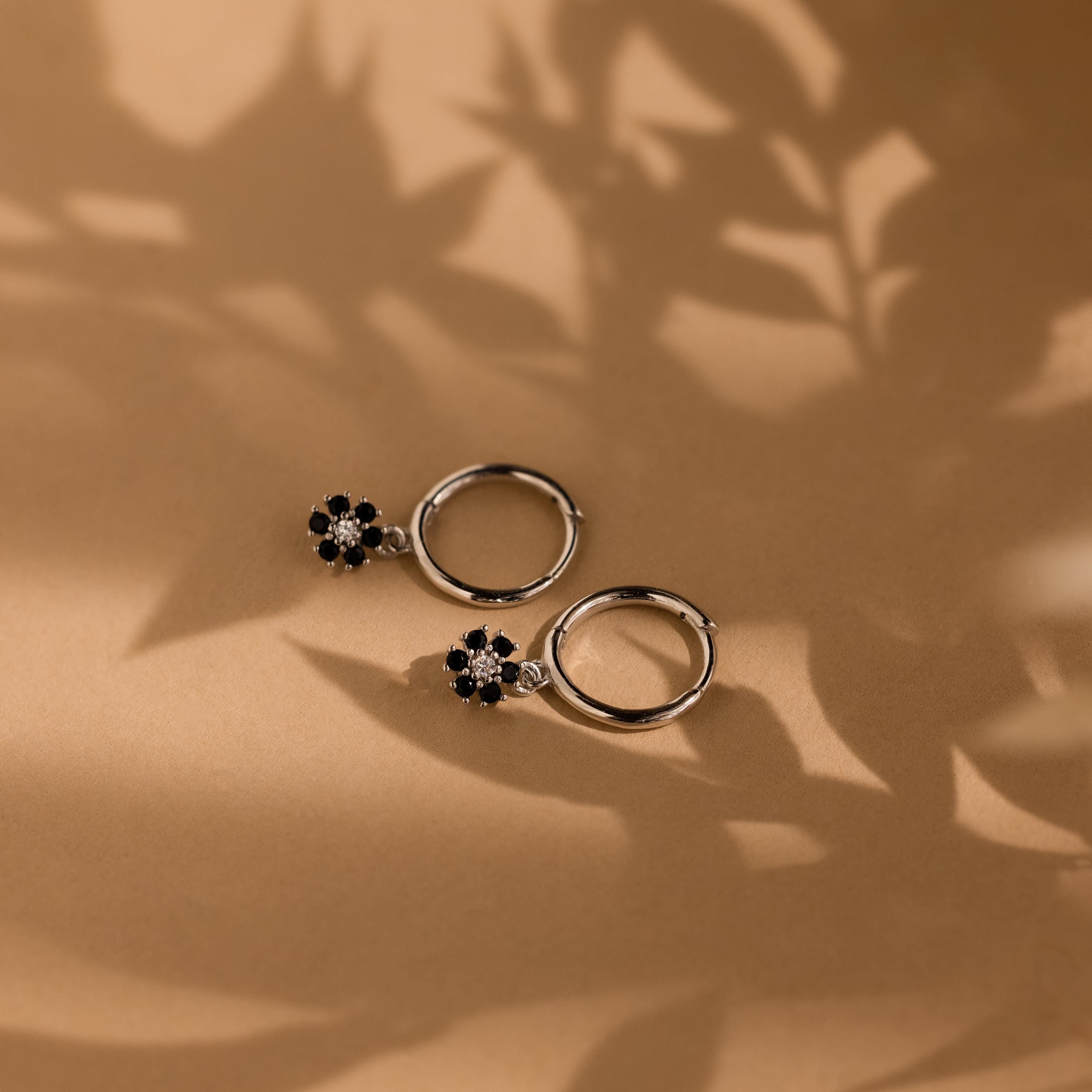 Two Onyx Flower Huggies, featuring silver hoops with black onyx flower charms, rest on a brown surface with leafy shadows, highlighting their elegant floral design.