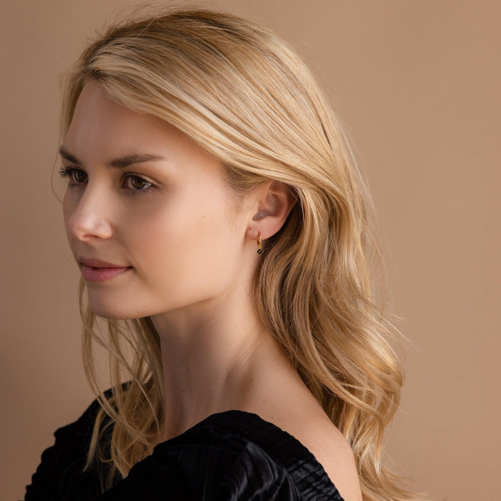 A woman with wavy blonde hair in a black top looks to the side against a beige background, highlighting the elegant Black Ivy Flower Huggies.