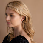 A woman with wavy blonde hair in a black top looks to the side against a beige background, highlighting the elegant Black Ivy Flower Huggies.