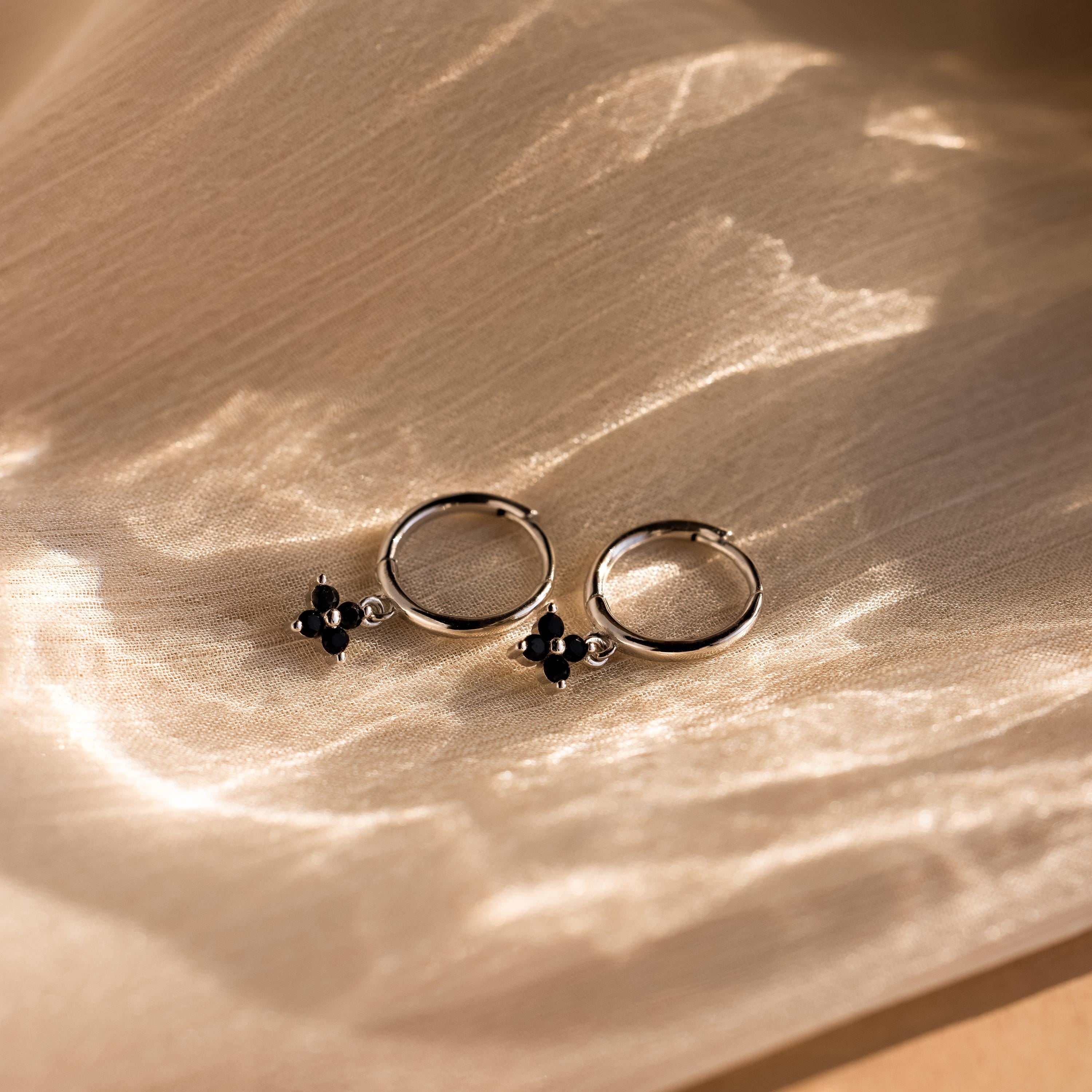 Black Ivy Flower Huggies: Small silver hoop earrings with black cross-shaped charms, shown on textured beige fabric.