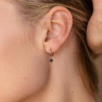 A close-up shows a woman's ear adorned with the elegant Black Ivy Flower Huggies—small silver hoop earrings featuring a black stone.