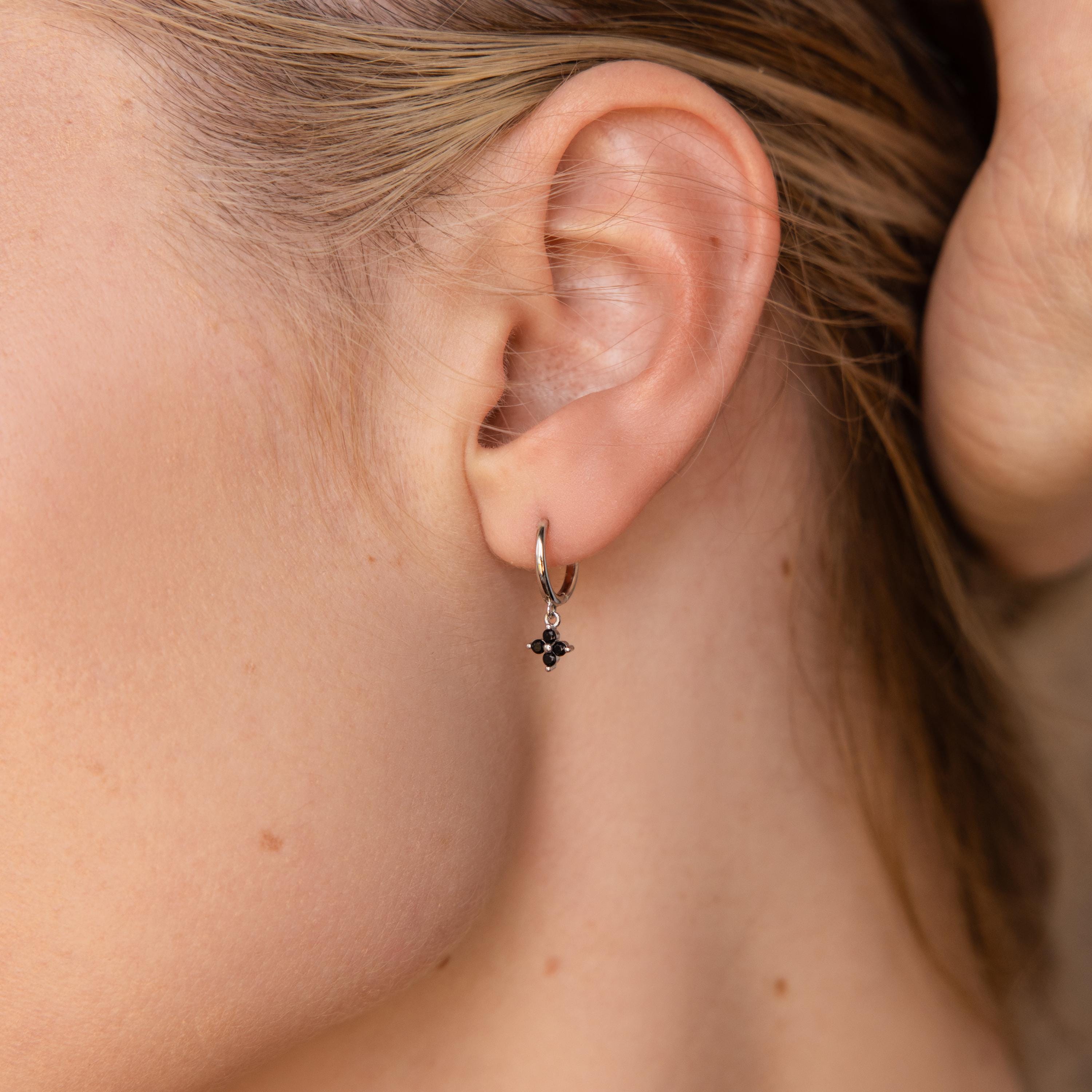 A close-up shows a woman's ear adorned with the elegant Black Ivy Flower Huggies—small silver hoop earrings featuring a black stone.