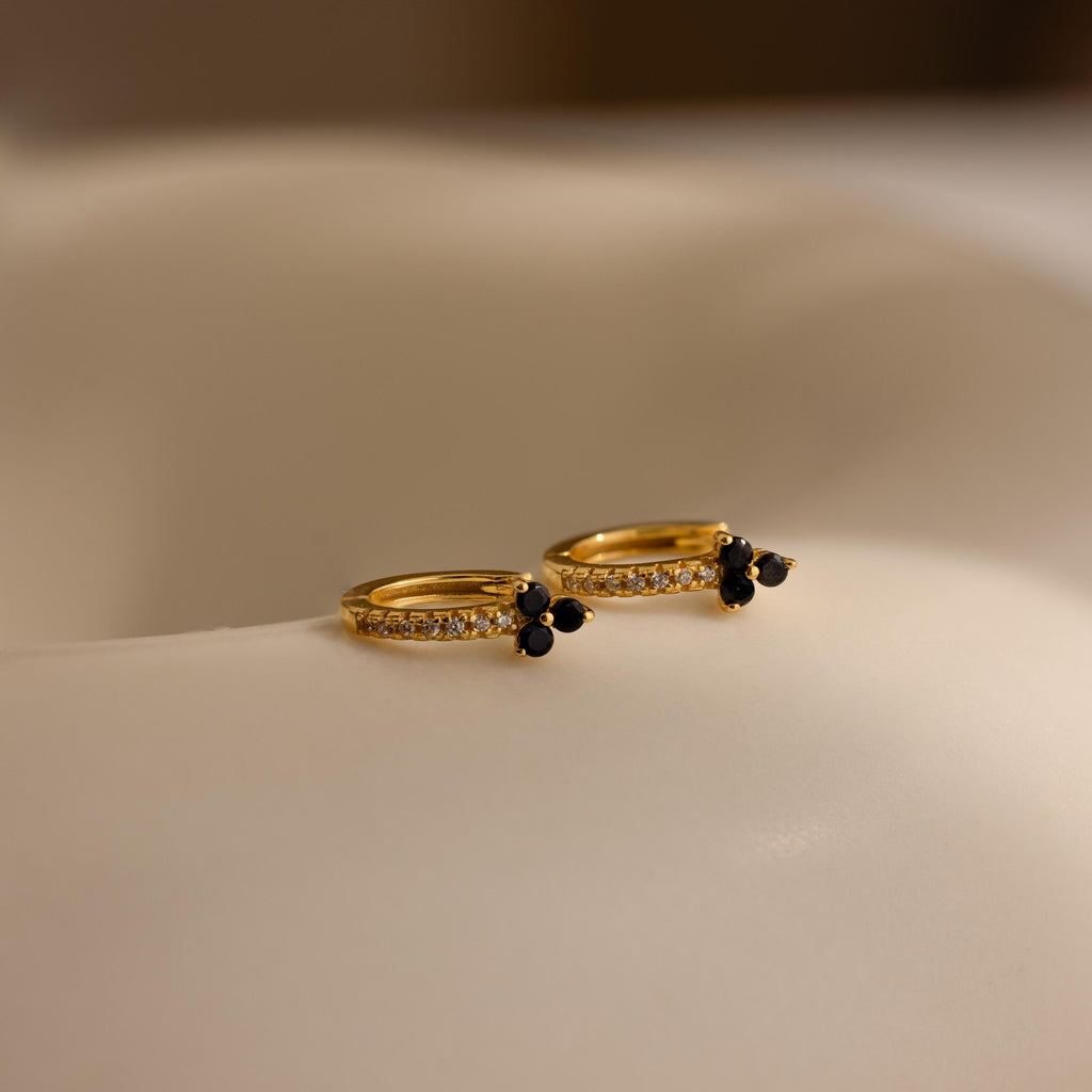 The Onyx Lily Huggies are gold hoops featuring small diamonds and black stones, elegantly showcased on a soft ivory surface.