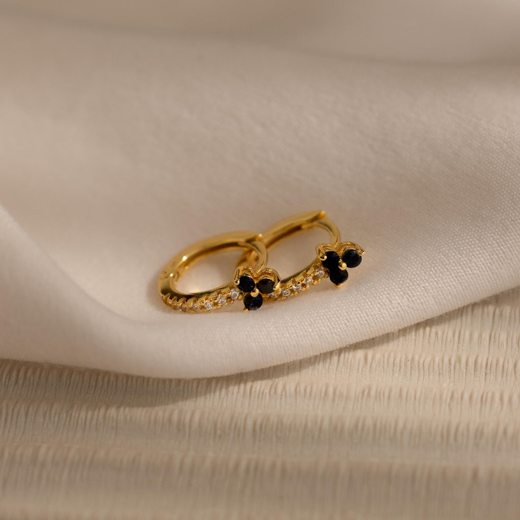 The Onyx Lily Huggies are gold hoop earrings adorned with small black stones and diamonds, shown on soft white fabric for an elegant, dainty appearance.