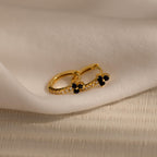 The Onyx Lily Huggies are gold hoop earrings adorned with small black stones and diamonds, shown on soft white fabric for an elegant, dainty appearance.