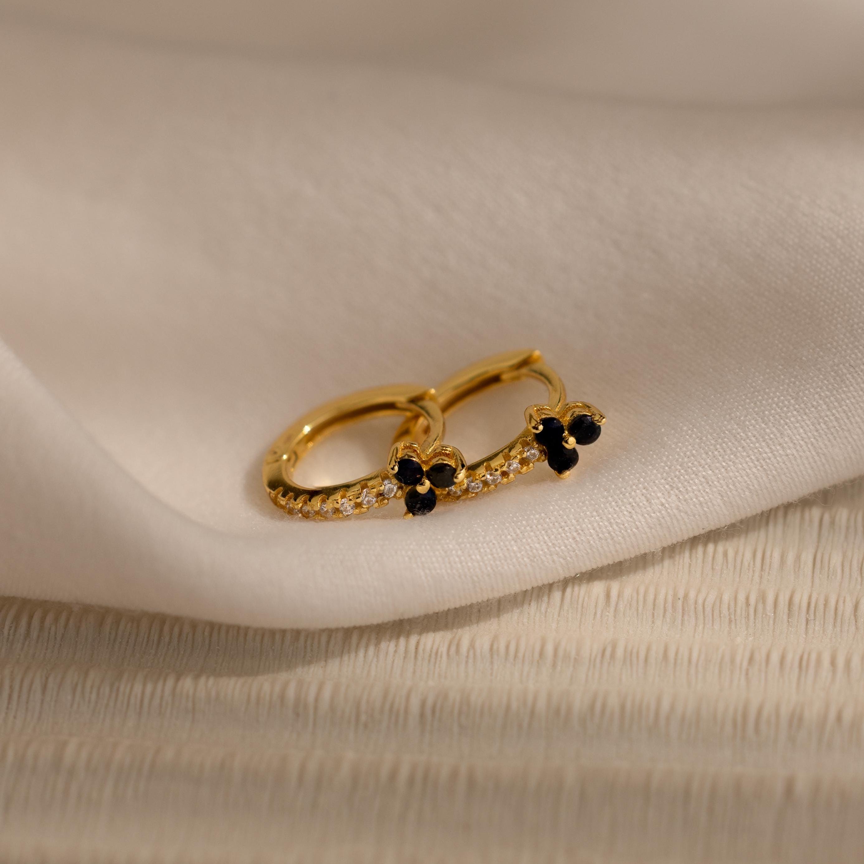 The Onyx Lily Huggies are gold hoop earrings adorned with small black stones and diamonds, shown on soft white fabric for an elegant, dainty appearance.