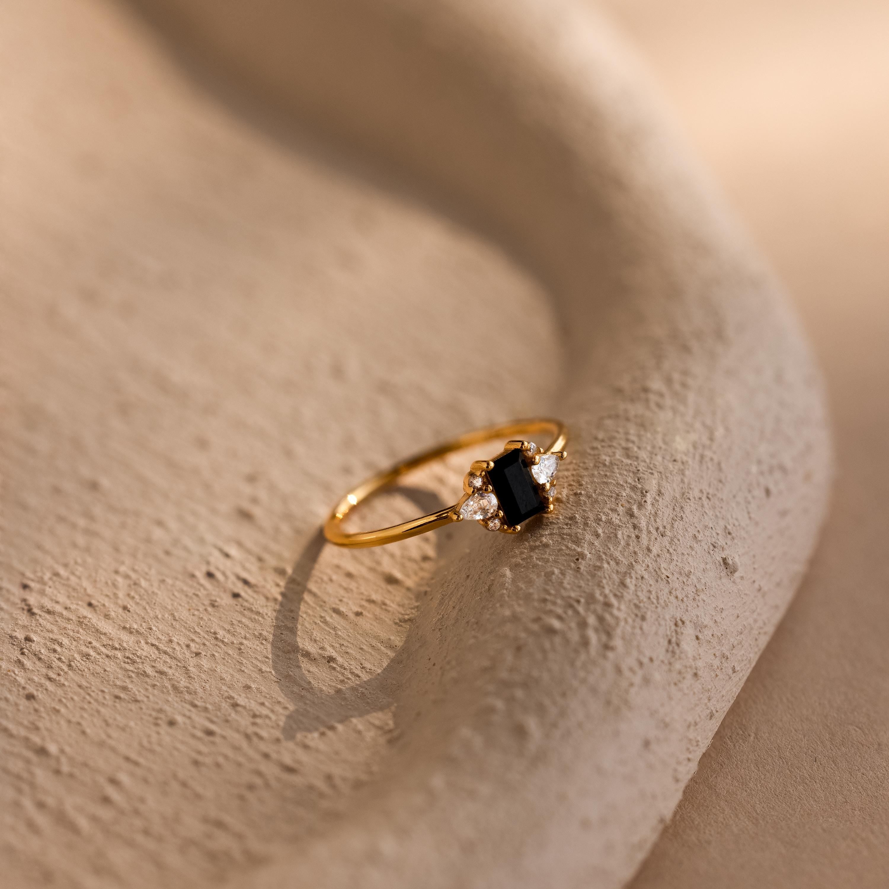 The Black Baguette Diamond Ring in 18K Gold features a rectangular black stone and clear gems, shown on a textured beige background—a bold vintage-inspired statement accessory.