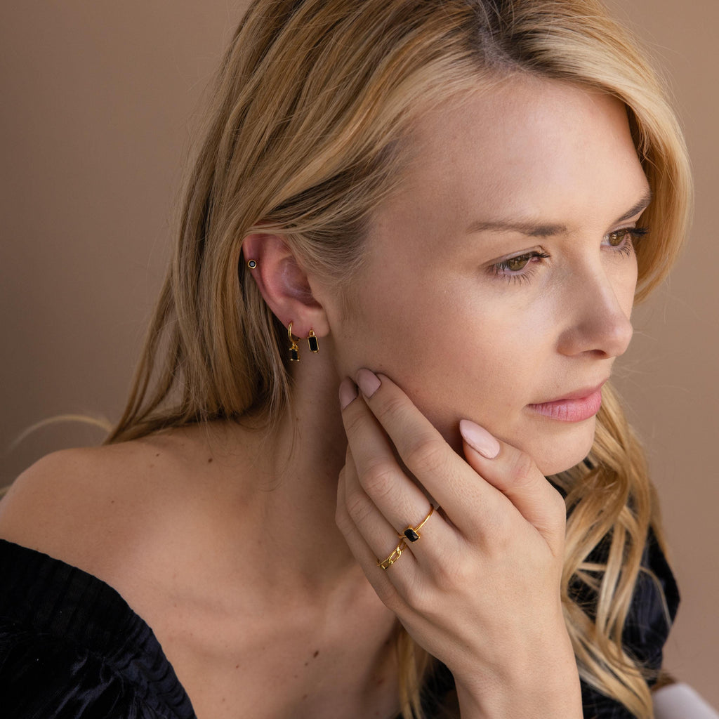 A blonde woman looks thoughtful, wearing gold jewelry including the Onyx Link Huggies and rings, with her hand gently touching her face.