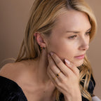 A blonde woman looks thoughtful, wearing gold jewelry including the Onyx Link Huggies and rings, with her hand gently touching her face.