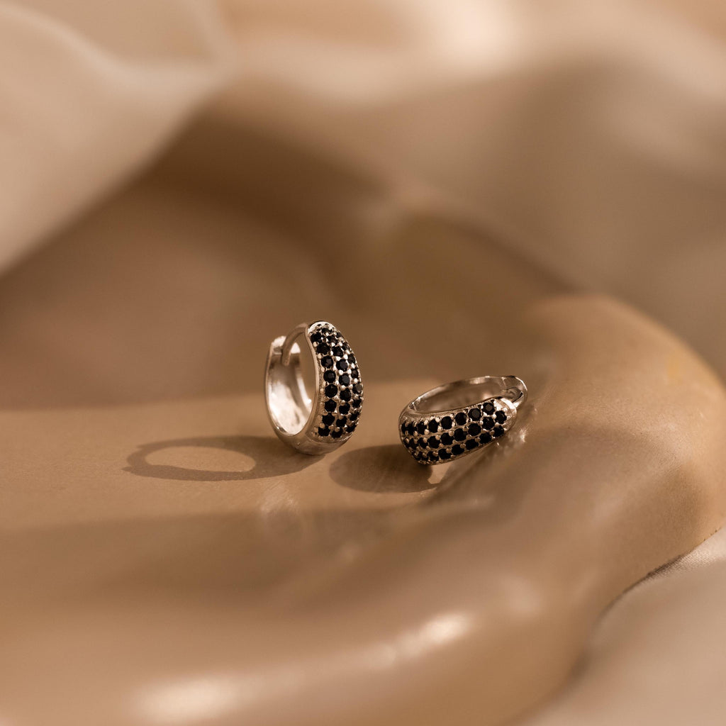 The Chunky Onyx Pave Huggies—sleek silver hoops set with pavé black onyx stones—rest on a smooth beige surface under soft lighting, offering a modern twist on classic black onyx earrings.