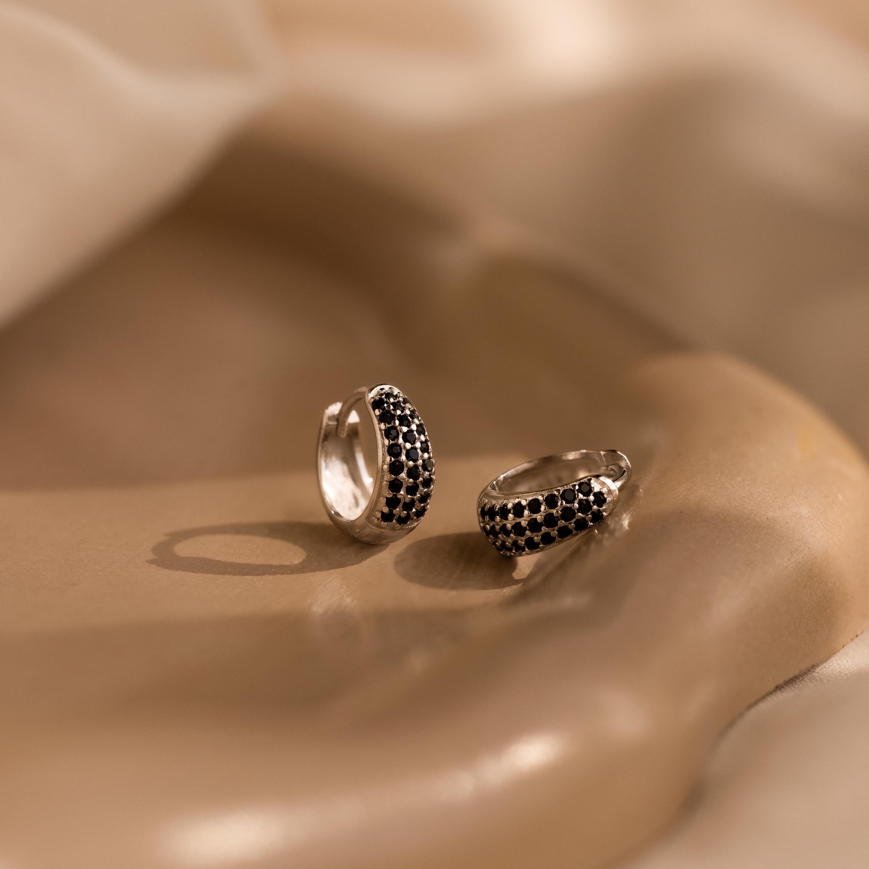The Chunky Onyx Pave Huggies—sleek silver hoops set with pavé black onyx stones—rest on a smooth beige surface under soft lighting, offering a modern twist on classic black onyx earrings.