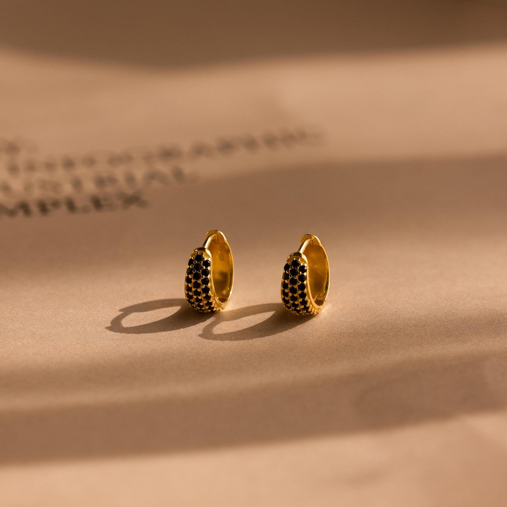 Chunky Onyx Pave Huggies—gold hoop earrings with black stones—cast bold shadows on a beige surface, blending modern style with classic elegance.