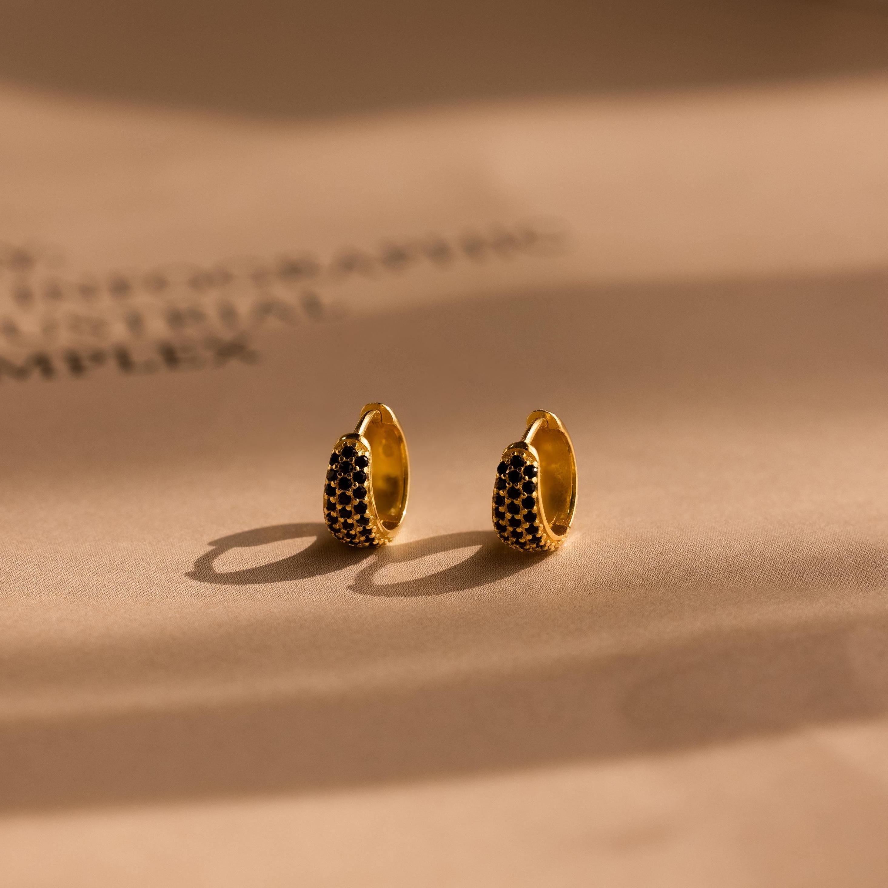 Chunky Onyx Pave Huggies—gold hoop earrings with black stones—cast bold shadows on a beige surface, blending modern style with classic elegance.
